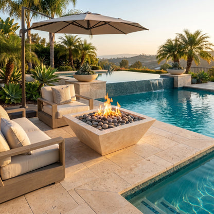 Outdoor patio with fire pit, chairs, and umbrella by a pool with palm trees in the background. Miletos Ivory Travertine Natural Stone Fire Pit Dimensions: 24" W x 24" L x 8" H