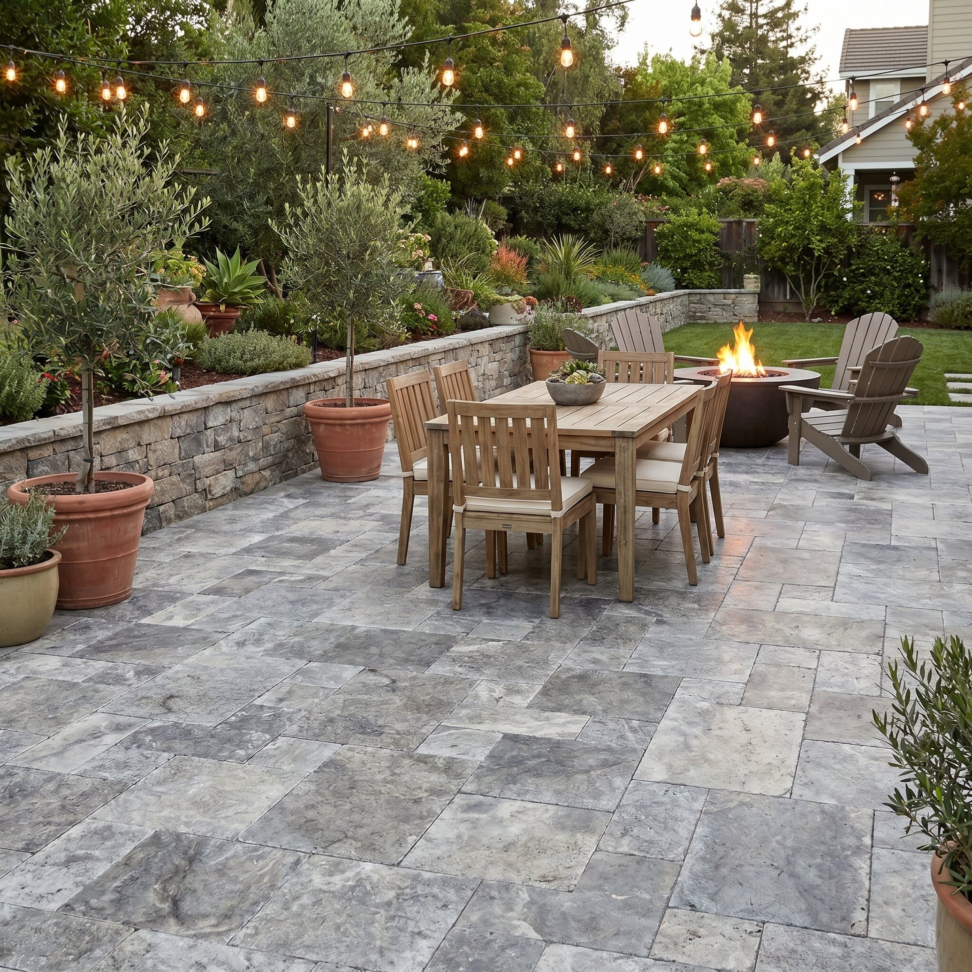 Outdoor patio with wooden furniture, fire pit, and string lights. Silver Travertine Tumbled Pavers
