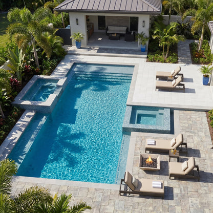A luxurious pool area with lounge chairs, a fire pit, and a modern house in the background. Silver Travertine Natural Stone Fire Pit – 24" x 24" x 8"