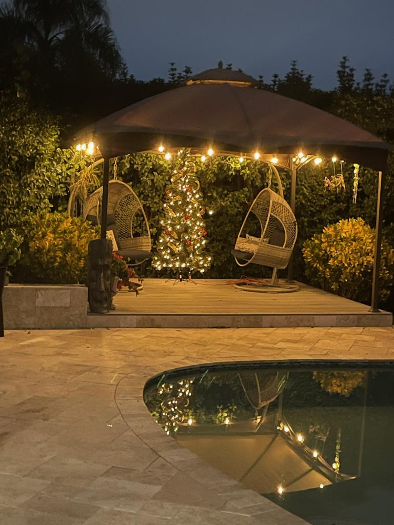 Gazebo with string lights, wicker chairs, and a Christmas tree by a pool at night. Aspendos Tumbled Travertine Pavers