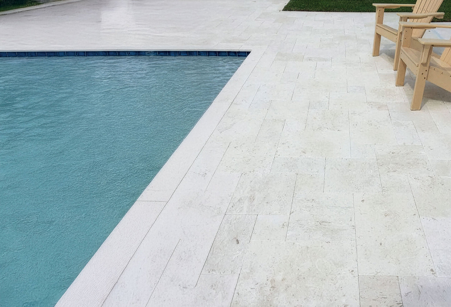 Poolside area with a pool and a wooden chair on a light-colored deck. Shell Stone White Tumbled Limestone Pavers